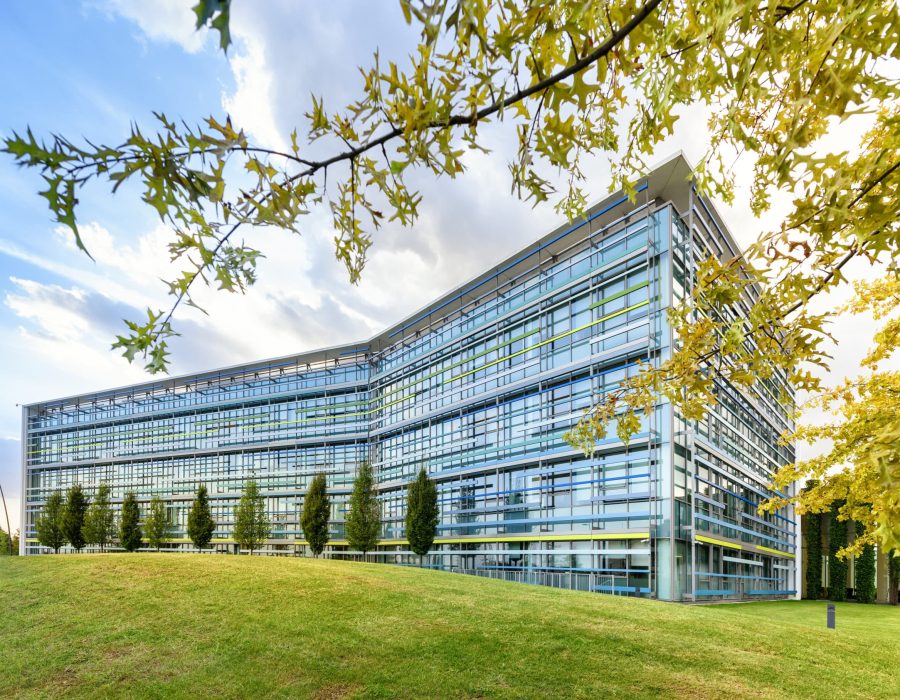 Exterior of a modern angular glass fronted office building in a spring landscape with green lawns and trees under a blue cloudy sky